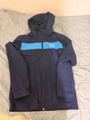 Marinblå vindjacka med huva - Snygg marinblå vindjacka från Under Armour med huva och dragkedja framtill. Jackan har en blå panel över bröstet med logga och är tillverkad i lätt, vindtätt material. Perfekt för blåsiga dagar och sportiga outfits.