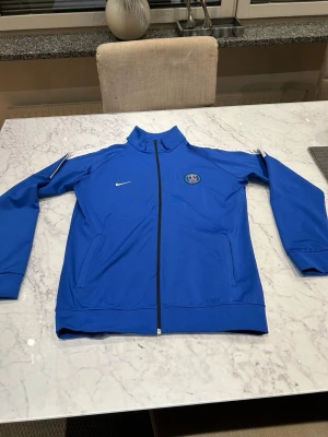 Blå Nike PSG träningsjacka - Snygg blå träningsjacka från Nike med PSG-logga på bröstet. Jackan har hel dragkedja, ståkrage och två fickor framtill. Perfekt för fotbollsträning eller chill. Materialet är troligtvis polyester och jackan har vita detaljer på axlarna.