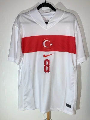 L. Äkta Turkiet 2024/2025 1st - Har använt max 5 gånger totalt 10 timmar Jag är en fotbolls tröja samlare i över 15år nu väljer jag och sälja den pågrund av min ps5 gick sönder för 2 dagar sen behöver köpa en ny därför väljer jag sälja den här en av några fotbolls tröjorVit fotbollströja från Nike med röd horisontell rand och Turkiets måne och stjärna på bröstet. Nummer 8 och Arda Güler tryckt på ryggen. Perfekt för dig som vill stötta Turkiets landslag. Tröjan har korta ärmar och V-ringning.