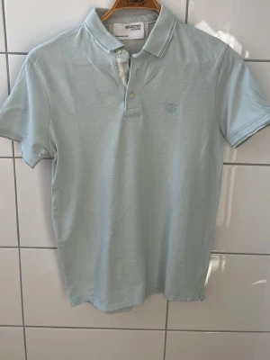 Ljusblå pikétröja från Selected Homme - Snygg ljusblå pikétröja från Selected Homme med klassisk krage och knappar framtill. Tröjan har korta ärmar och diskret broderad logga på bröstet. Perfekt till jeans eller shorts för en clean och fräsch look. Har liten fläck i slutet av tröjan