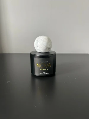 Nova Naiara X parfym 50 ml - Elegant parfymflaska i svart med guldfärgad text och en unik, rund vit marmorerad kork. Flaskan rymmer 50 ml och har en modern, stilren design som sticker ut på parfymhyllan. Perfekt för dig som gillar exklusiva dofter och snygg inredningsdetalj.