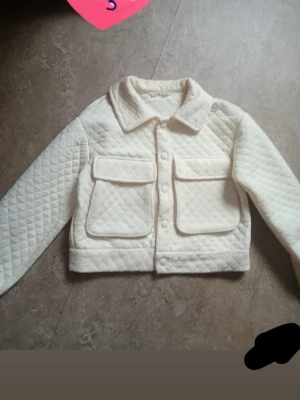Vit quiltad cardiganjacka - Quiltad cardiganjacka i offwhite med stora fickor framtill och knappar längs hela framsidan. Jackan har krage och långa ärmar, samt ett rutmönster i tyget som ger en trendig och mysig känsla. Perfekt för lager på lager och en clean look.
