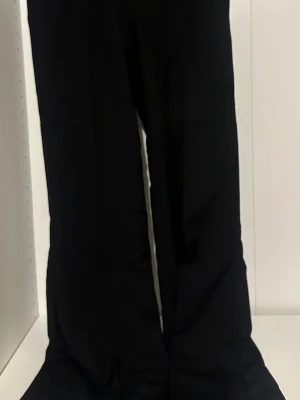 Svarta vida kostymbyxor - Säljer ett par svarta kostymbyxor med hög midja och vida ben. Byxorna har en klassisk bootcut passform och är perfekta för en clean och stilren look. Materialet ser ut att vara mjukt och följsamt, vilket ger en snygg siluett.