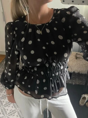 Svart prickig blus med resårmidja - Säljer en svart blus med vita stora prickar och transparenta långärmar. Blusen har resår i midjan som ger en snygg siluett och volang nedtill. Materialet är tunt och luftig, perfekt för en trendig look.