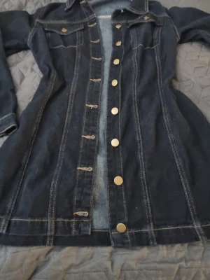 Mörkblå jeansklänning med knappar - Snygg mörkblå jeansklänning med långa ärmar och guldiga knappar framtill. Klänningen har klassisk skjortkrage, bröstfickor med lock och markerade sömmar som ger en cool denimlook. Perfekt för dig som gillar en avslappnad men trendig stil. Oanvänd I storlek M. 