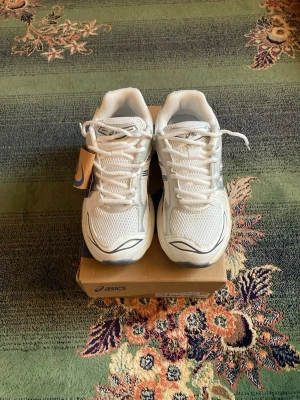 Vita Asics sneakers med silverdetaljer - Snygga vita sneakers från Asics med silver- och grå detaljer. Skorna har en sportig design med meshpaneler för bra ventilation och robust sula för extra komfort. Perfekta för dig som gillar en clean och fräsch look till vardagsoutfiten.