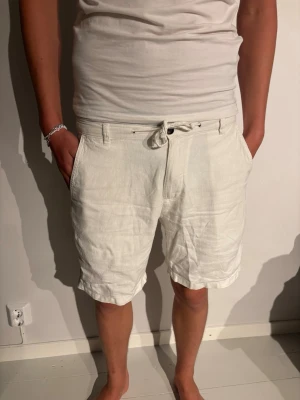 Vita linneshorts med snörning - Snygga vita shorts i linne med snörning i midjan och fickor både fram och bak. Klassisk passform, Perfekta för varma dagar och ger en fräsch look.