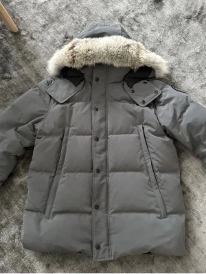 Grå dunjacka med fuskpäls - Snygg grå dunjacka med stor huva och fuskpälsdetalj. Jackan har två stora snedställda fickor framtill, knäppning med tryckknappar och dragkedja samt vadderad design för extra värme. Perfekt för kalla vinterdagar.