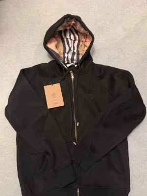 Svart ziphoodie från Burberry - Svart ziphoodie från Burberry. Perfekt för en stilren och trendig look.                             Storlek L men passar även i M 