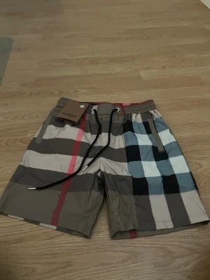 Burberry swimware - Snygga badshorts med klassiskt rutigt mönster i beige, svart, vit, röd och blå. Shortsen har elastisk midja med svart snörning och sidofickor. Perfekta för stranden eller poolen och har en loose passform för skön känsla.