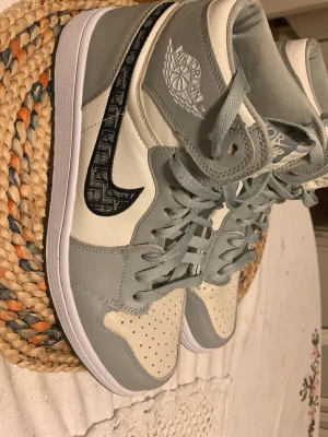 Gråvita höga sneakers med svart swoosh - Stilrena höga sneakers i grått och vitt med svart mönstrad swoosh på sidan. Skorna har perforerad tå, klassisk snörning och Air Jordan-logga på sidan. Tillverkade i läder med vit sula och grå detaljer. Perfekt för dig som gillar streetwear och sportig vibe.
