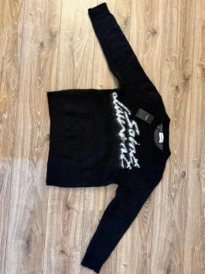 Svart stickad tröja Saint Laurent - Svart stickad tröja från Saint Laurent med vit logotyp framtill i en cool, sprayliknande stil. Tröjan har rund halsringning och långa ärmar. Mjuk och fluffig känsla, perfekt för att lyfta din outfit.