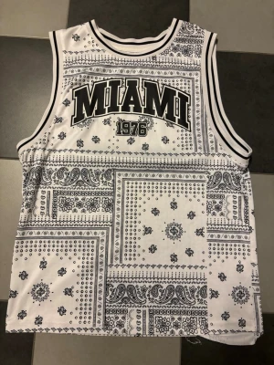 Vit och svart Miami linnetopp - Säljer en vit och svart linnetopp med bandanamönster och texten 'MIAMI 1976' framtill. Toppen är ärmlös med sportiga ränder vid hals och ärmhål, perfekt för en avslappnad och trendig look.