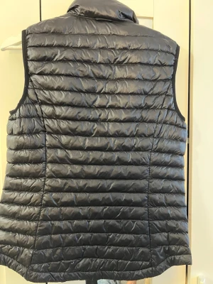 Svart lättviktsdunväst - Snygg svart dunväst med quiltad design och hög krage. Västen är lätt och smidig, perfekt för lager-på-lager och har en diskret glans i materialet. Dragkedja framtill och smal passform som ger en modern look.
