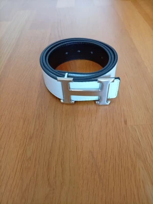 Vändbart vitt och svart skärp med H-spänne - Snyggt vändbart skärp i vitt och svart med ett stort H-format spänne i silverfärg. Bältet är gjort i skinn och har en stilren, klassisk look som passar till många outfits. Perfekt accessoar för dig som vill ha något både trendigt och tidlöst.