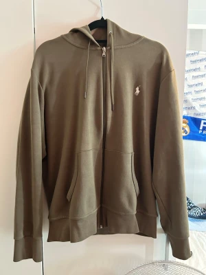 Olivgrön Ralph Lauren hoodie med dragkedja - Säljer en olivgrön hoodie från Polo Ralph Lauren med dragkedja framtill, två fickor och justerbar huva med snören. Klassisk logga broderad på bröstet. Perfekt för chill dagar och enkel att matcha med jeans eller sweatpants.