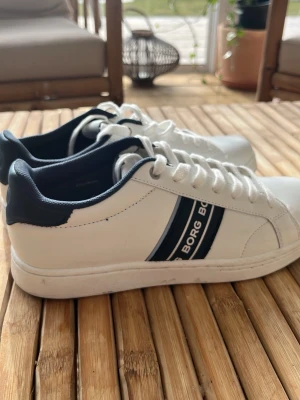 Vita Björn Borg sneakers med blå detaljer - Snygga vita sneakers från Björn Borg med mörkblå detaljer och två ränder på sidan. Skorna har klassisk låg profil, vita snören och en stilren design som passar till det mesta. Tillverkade i slätt syntetmaterial med bekväm innersula.