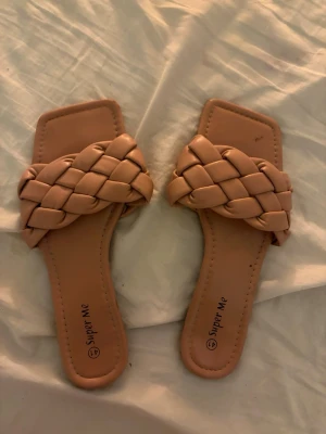 Beige flätade slip-in sandaler - Säljer ett par trendiga slip-in sandaler från Super Me i beige färg. De har en bred, flätad rem över foten och fyrkantig tå. Tillverkade i ett mjukt, konstläderliknande material som känns bekvämt mot huden. Perfekta för en avslappnad och stilren look.