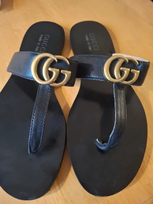 Svarta sandaler från Gucci med GG-logo - Snygga svarta sandaler från Gucci i läder med klassisk GG-logo i guld framtill. Tårem-design och stilren look som passar perfekt till sommaren. Kommer med originalpåse och kontrollkort från Gucci.