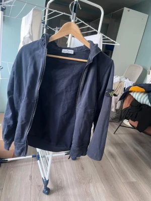 Mörkblå hoodie från Stone Island - Säljer en mörkblå hoodie från Stone Island Junior med dragkedja framtill och klassisk logga på vänster ärm. Hoodien har huva och två fickor framtill. Tillverkad i mjukt bomullsmaterial som känns skönt mot huden. Perfekt för en avslappnad och stilren look.