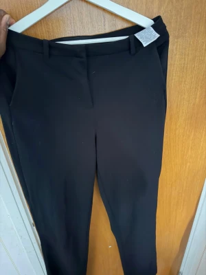 Svarta kostymbyxor från Vero Moda - Stilrena svarta kostymbyxor från Vero Moda med smal passform och klassiska bälteshällor. Byxorna har diskreta sidofickor och stängs med dragkedja och hake. Perfekta för en clean och modern look. Säljes pågrund av ingen användning.
