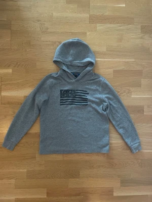Grå hoodie från Polo Ralph Lauren - Snygg grå hoodie från Polo Ralph Lauren med svart flaggtryck på bröstet. Tillverkad i mjukt, ribbat material och har en klassisk huva. Perfekt för en avslappnad och cool stil.