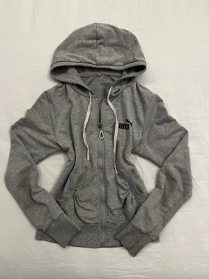 Vintage Puma Grå Zip Huvtröja | Y2K | XS/S - Så vacker grå zip huvtröja från Puma som passar perfekt till träning eller vardag. Den ger av en Y2K vibe med tryck på ryggen, fina detaljer på dragkedjan och dekorativa skrynkor vid fickan. Väldigt bekväm, i 95% bomull. Bra skick. Pris kan diskuteras vid snabb affär. 