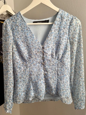 Blommig blå blus från Vero Moda - Supersöt blommig blus från Vero Moda i ljusblått med små bruna och vita blommor. Blusen har v-ringning, knappar framtill och långa ärmar. Tunt, lätt material som känns luftigt och perfekt till våren eller sommaren.