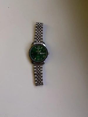 klocka med grön urtavla - Stilren klocka med grön urtavla och romerska siffror. Armbandet är i silverfärgad metall med klassisk länkdesign. Klockan har en rund form och tydliga detaljer på urtavlan. Perfekt accessoar för dig som vill sticka ut.