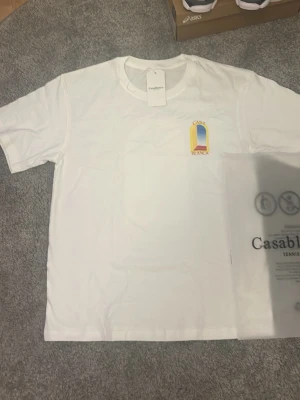 Vit t-shirt från Casablanca - Säljer en fräsch vit t-shirt från Casablanca med ett färgglatt tryck på bröstet. T-shirten har rund hals och korta ärmar, tillverkad i mjuk bomull. Perfekt för en clean och stilren look.