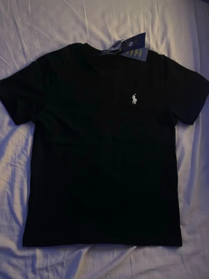 Svart t-shirt från Polo Ralph Lauren - Strl S - Klassisk svart t-shirt från Polo Ralph Lauren med rund halsringning och ikonisk vit broderad logga på bröstet. Tillverkad i mjuk bomull för en skön känsla och perfekt passform. Enkel och stilren design som funkar till allt.