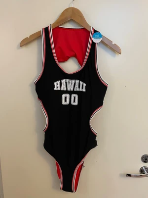 Svart sportig baddräkt Hawaii 00, ny storlek L  - Sportig svart baddräkt med vita och röda detaljer samt texten 'HAWAII 00' framtill. Kontrasterande röd insida och vita/röda kanter ger en cool look. Modellen har en öppen rygg och är perfekt för strand eller pool. Stl L