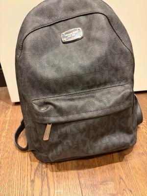 Svart ryggsäck från Michael Kors - Snygg svart ryggsäck från Michael Kors med diskret logomönster över hela väskan. Väskan har justerbara axelband, en stor ytterficka med dragkedja och metallplatta med logga framtill. Tillverkad i tåligt konstläder som ger en lyxig känsla.