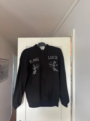 Svart Zip från Bling Lucid  - Svart Zip från Bling Lucid. På bröstet står det 'BLING LUCID i glittriga stenar tillsammans med två änglamotiv i samma stil. Hoodien har fickor framtill och ribbade muddar vid ärmar och nederkant. Perfekt för dig som gillar statement-plagg.