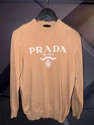 Prada tröja - Snygg beige stickad tröja från Prada med vit logga och texten 'PRADA MILANO' framtill. Tröjan har rund halsringning, ribbade muddar och en klassisk passform. Perfekt för dig som gillar exklusiva och stilrena plagg.