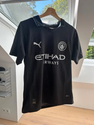 Svart Manchester City Tröja - Snygg svart Manchester City matchtröja från Puma med vit logga och Etihad Airways-tryck på bröstet. Tröjan har korta ärmar, krage och är tillverkad i ett lätt, snabbtorkande material. Perfekt för fotboll eller träning.
