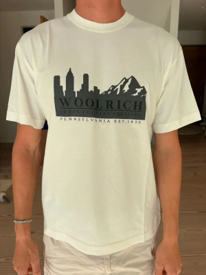 Vit Woolrich t-shirt med tryck - Helt ny Vit t-shirt från Woolrich med svart tryck av stadssil och berg på bröstet med lapparna kvar. Klassisk rund hals och normal passform. Tillverkad i mjuk bomull som känns skön mot huden. Nypris 766 kr.