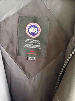 Svart parkas från Canada Goose XS - Säljer en svart parkas från Canada Goose i storlek XS. Jackan har huva, långa ärmar och klassisk Canada Goose-logga på ärmen. Tillverkad i slitstarkt material och fodrad för extra värme. Perfekt för kalla dagar och riktigt snygg streetstil. Jackan är den långa modellen. 