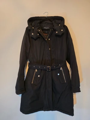 Hollies Stockholm svart dunparkas med bälte – stl 40  - 🖤 Stilren svart dunparkas från Hollies Stockholm i storlek 40. Varm och klassisk modell med avtagbart midjebälte, huva och fickor med dragkedja. Jackan har 80 % dun och 20 % fjäder och kommer även med en löstagbar stickad grå ullkrage som ger extra värme under kalla dagar. ✨ Sparsamt använd som finjacka under två säsonger och i mycket fint skick. Dunet är fortfarande jämnt fördelat, puffigt och fint utan klumpar. Endast kemtvättad. 🔖 Storlek: 40 🧵 Fyllning: 80 % dun, 20 % fjäder 🏷️ Nypris: 3 995 