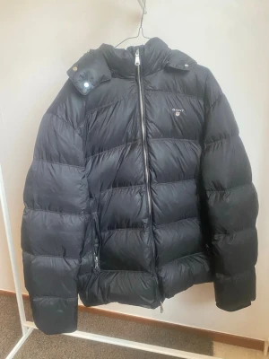 Svart pufferjacka från Gant - Snygg svart pufferjacka från Gant med huva och dragkedja framtill. Jackan har två sidofickor med dragkedja och Gant-logga på bröstet. Perfekt för kalla vinterdagar tack vare den tjocka vadderingen och den höga kragen.