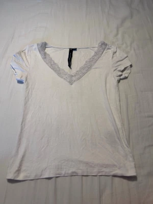 Vit topp med spetsdetalj och v-ringning - Snygg vit topp med v-ringad hals och spetsdetalj runt ringningen. Kortärmad och i mjukt material som känns skönt mot huden. Perfekt att styla med jeans eller kjol för en fräsch look.