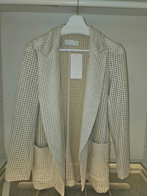 Beige perforerad kavaj med fickor - Trendig beige kavaj med perforerat mönster och två stora fickor framtill. Kavajen har klassisk krage och är tillverkad i ett lätt material som ger en luftig känsla. Perfekt för dig som vill ha en unik och stilren look.