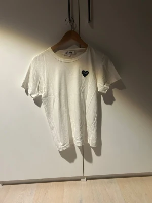 Vit Comme des Garçons Play t-shirt - Säljer en vit t-shirt från Comme des Garçons Play med det ikoniska svarta hjärtat med ögon broderat på bröstet. T-shirten har rund hals och korta ärmar, tillverkad i mjuk bomull för en skön och avslappnad look.