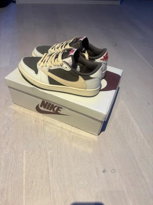 Nike Air Jordan 1 Low Travis Scott - Nike Air Jordan 1 Low Travis Scott med beige och mörkgrå paneler, off-white swoosh och röda detaljer på hälen. Skorna har en låg silhuett, klassisk snörning och är tillverkade i läder med textildetaljer. Perfekta för dig som gillar streetwear och unika sneakers. Aldrig använda bara provade på