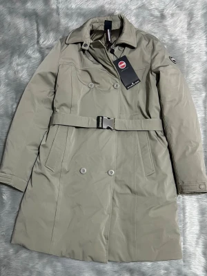 COLMAR ORIGINALS PADDED WATERPROOF FABRIC JACKET - Stilren beige trenchcoat från Colmar med bälte i midjan och dubbelknäppning framtill. Jackan har klassisk krage, två sidofickor och ett diskret Colmar-märke på ärmen. Materialet är slätt och vattenavvisande, perfekt för blåsiga eller kalla dagar. Ordinarie pris är 8799kr