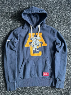 Mörkblå Superdry hoodie med tryck - Snygg mörkblå hoodie från Superdry med stor gul bokstavs- och kattapplikation på bröstet. Har vit brodyr på ärmen, vita dragsnören och känguruficka framtill. Perfekt för dig som gillar streetwear och vill sticka ut lite extra.