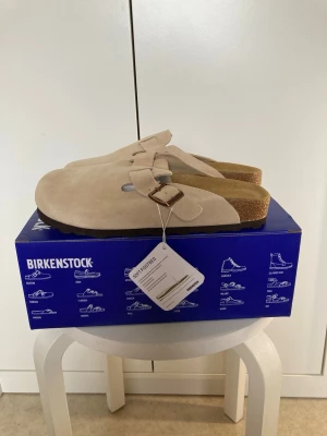 Beige Birkenstock tofflor mocka - Säljer ett par snygga beige Birkenstock tofflor i mocka med justerbart spänne i metall. Klassisk slip-in modell med mjuk innersula och korksula för extra komfort. Perfekta för dig som gillar stilrena och bekväma skor.