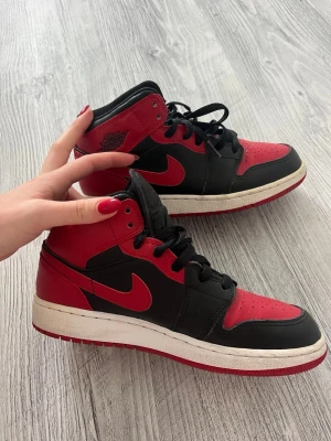 Nike Air Jordan 1 svart/röd sneakers - Säljer ett par klassiska Nike Air Jordan 1 sneakers i svart och rött med vita sulor. Skorna har högt skaft, svart snörning och ikoniska swoosh-loggan på sidan. Tillverkade i läder med snygga detaljer och retrovibe. Perfekta för dig som gillar streetwear och vill sticka ut.