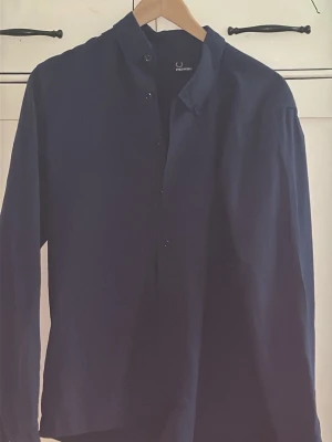 Mörkblå skjorta med knappar - Stilren mörkblå skjorta från Fred Perry med klassisk krage och dolda knappar framtill. Skjortan har lång ärm och rak passform, perfekt för en clean och enkel look. Tillverkad i ett mjukt material som känns bekvämt mot huden.