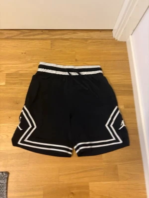 Svarta shorts med vita detaljer - Säljer ett par svarta shorts med vita ränder och snörning i midjan. Shortsen har elastisk midja och sportig look med grafiska linjer längs benen. Perfekta för dig som gillar en avslappnad och trendig stil.